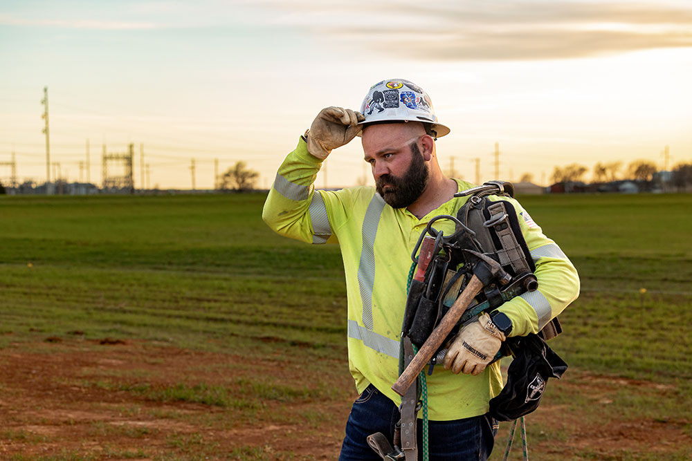 Un liniero, Kevin Walker, con casco y camisa de seguridad amarilla, se seca la frente mientras sostiene un cinturón de herramientas sobre su hombro en un campo de hierba al atardecer.